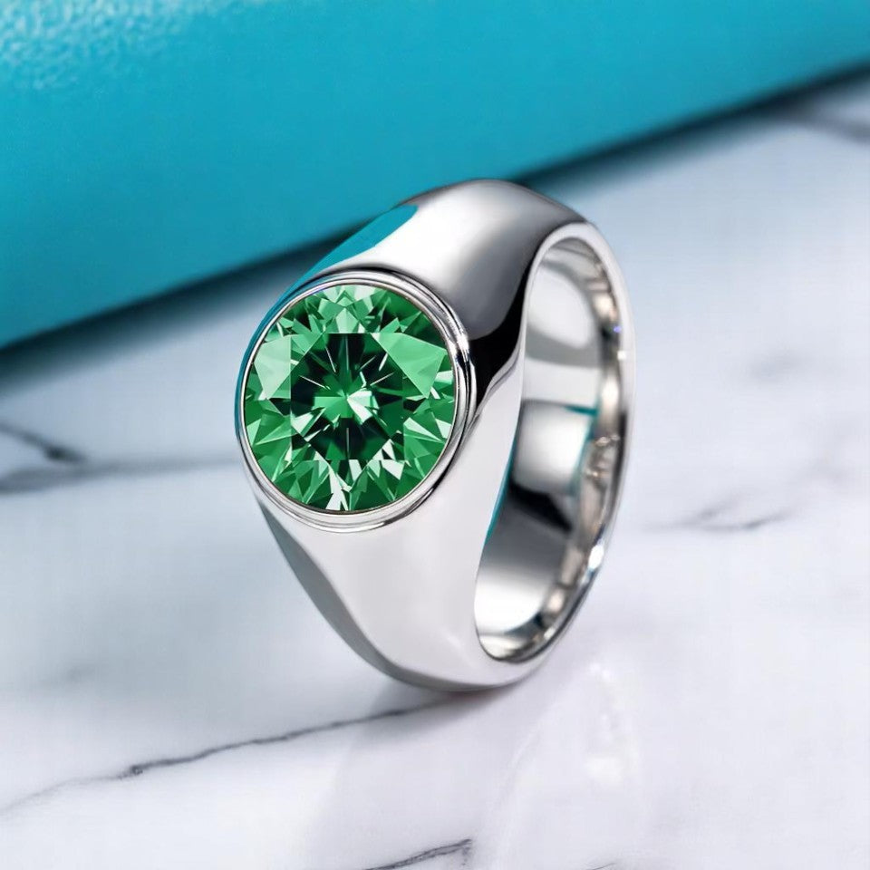 A rhodium-plated men's ring with a large round-cut emerald gemstone set in a silver band, displayed on a marble surface with a blue object in the background.